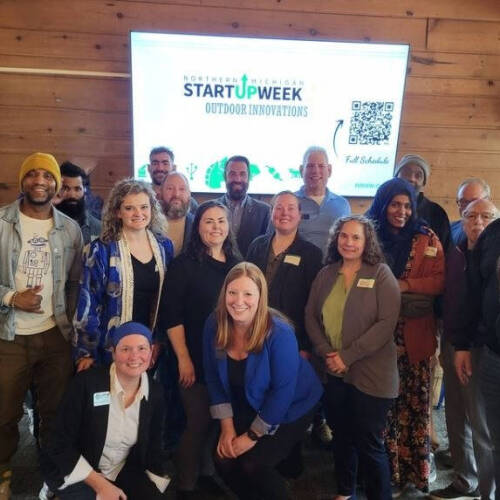 A group of people posing indoors with a screen displaying "Northern Michigan Startup Week: Outdoor Innovations" in the background.